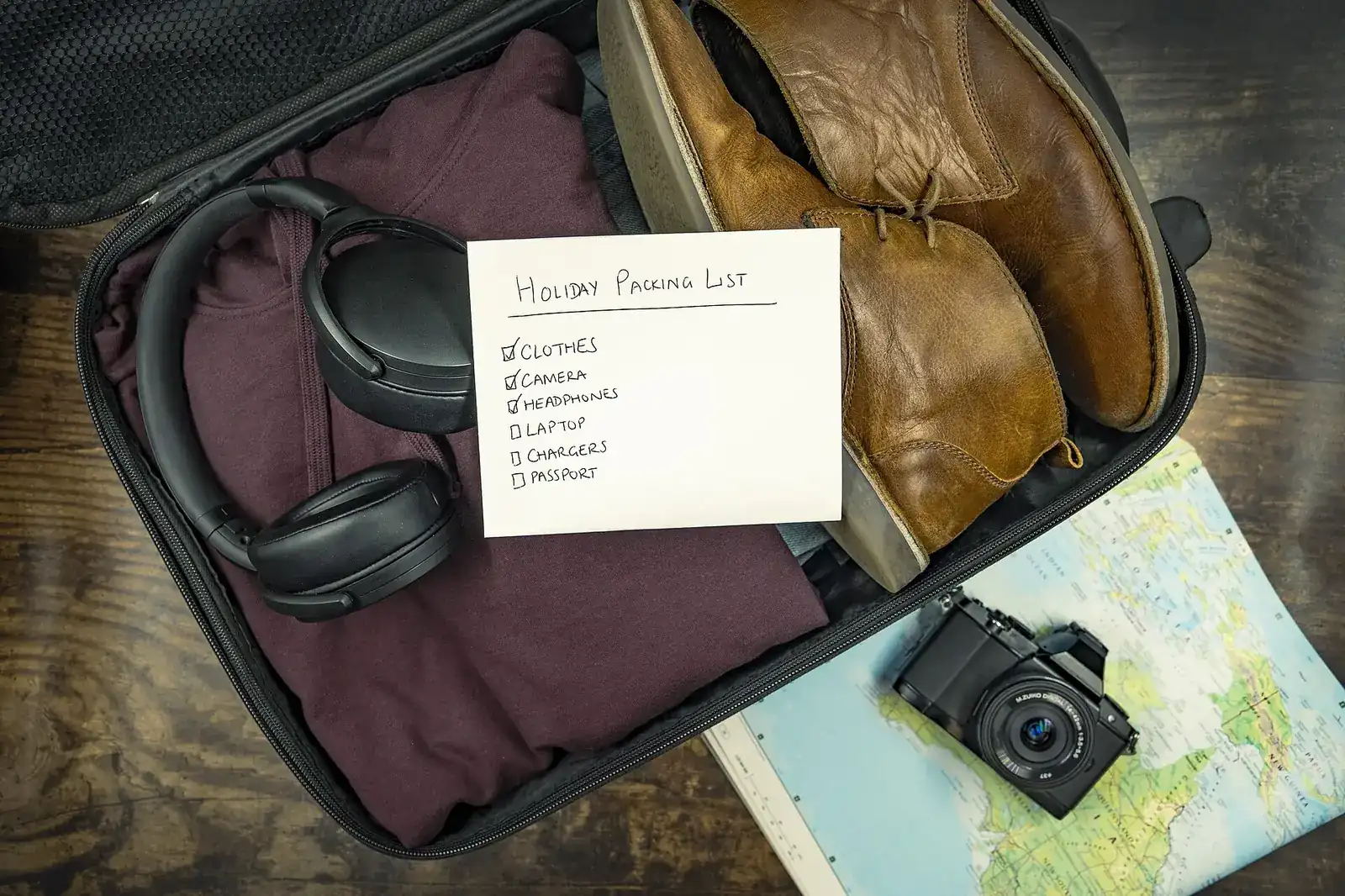 Bolsa de mano abierta con ropa, auriculares, una cámara, un mapa y una lista de equipaje escrita a mano