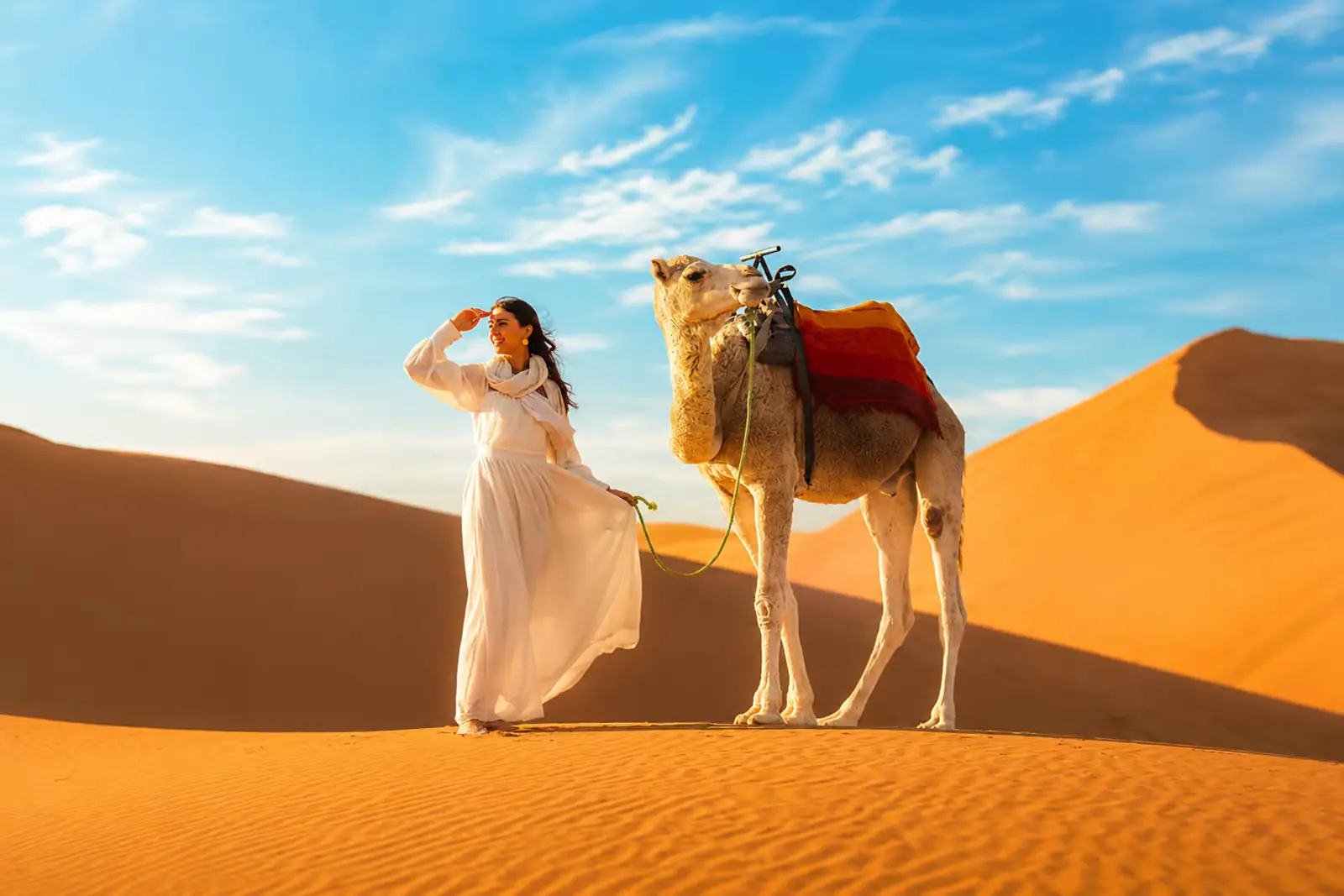 Mujer con vestido blanco junto a un camello en las dunas doradas de Merzouga