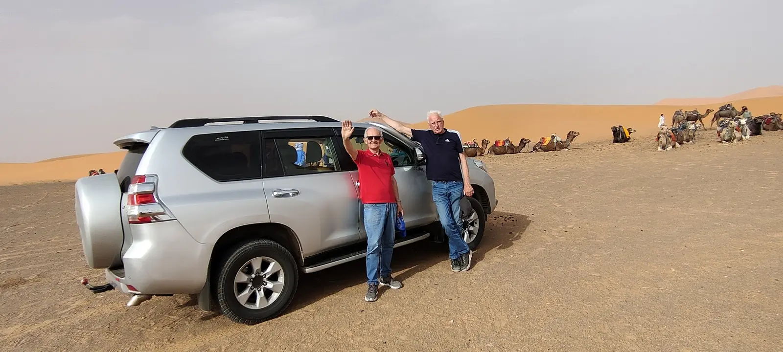 Two travellers waving beside a 4x4 on the desert in June