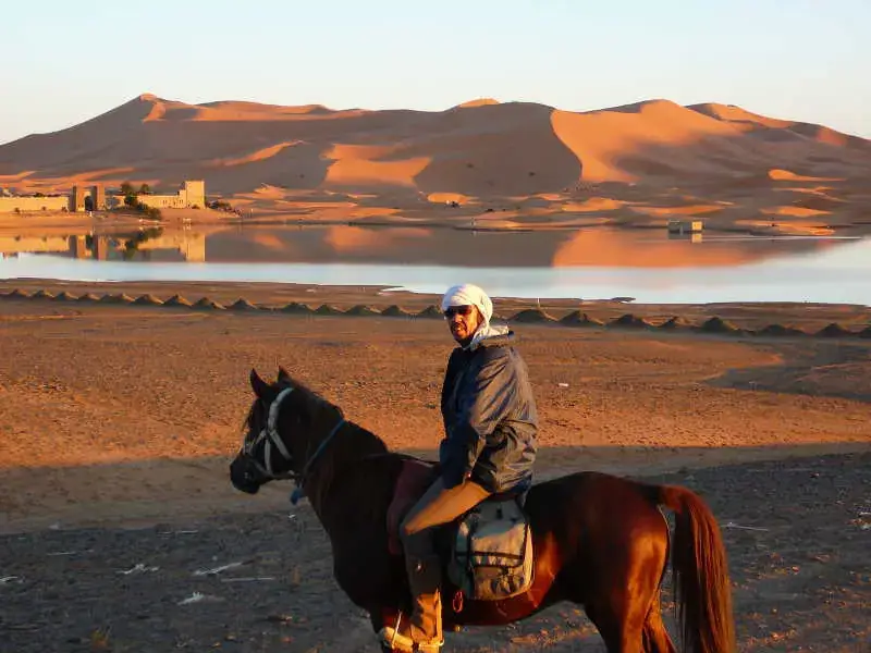 Rider on a horse beside a desert lake with dunes behind